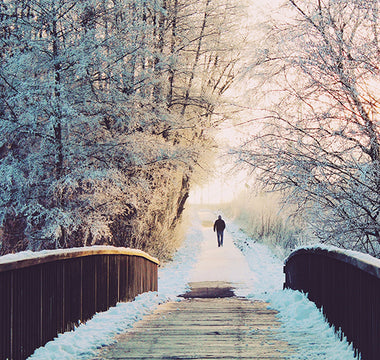 A person running or walking on the other side of a wooden bridge in a snowy landscape during sunset