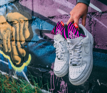 A hand holding a pair of white Nike sneakers with pink SmellWell inserts in them infront of a graffiti wall 