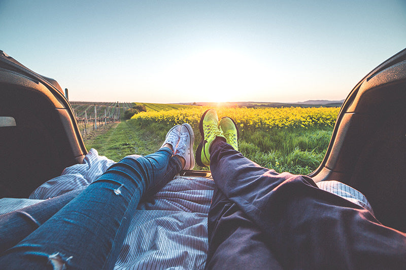 Two peoples pair of feet sticking out of the back of a car with a ciew over a green and yellow field in the sun