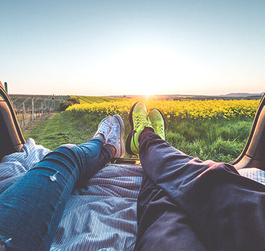 Two peoples pair of feet sticking out of the back of a car with a ciew over a green and yellow field in the sun