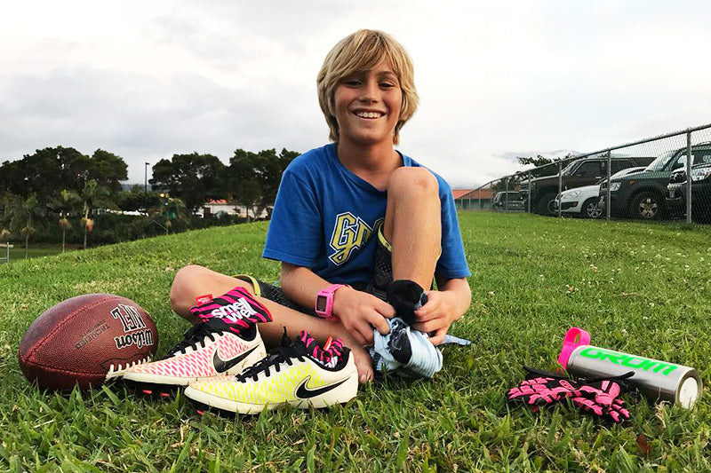 Smiling boy sitting on a grass field with a ball and a pair of Nike Football Shoes with SmellWell inserts in them.
