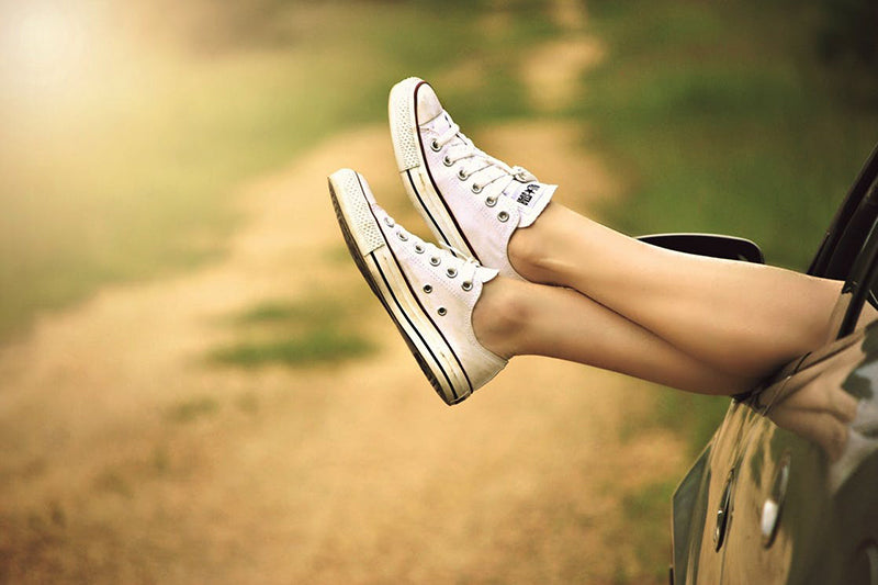 A pair of legs, with feet wearing white Converse sneakers, sticking out relaxed from a car window in summer time