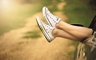 A pair of legs, with feet wearing white Converse sneakers, sticking out relaxed from a car window in summer time