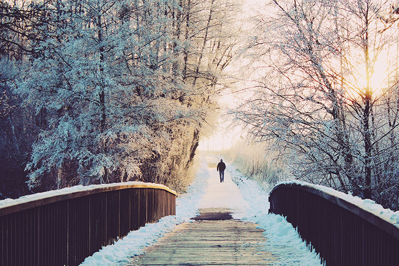 A person running or walking on the other side of a wooden bridge in a snowy landscape during sunset