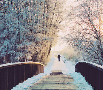 A person running or walking on the other side of a wooden bridge in a snowy landscape during sunset