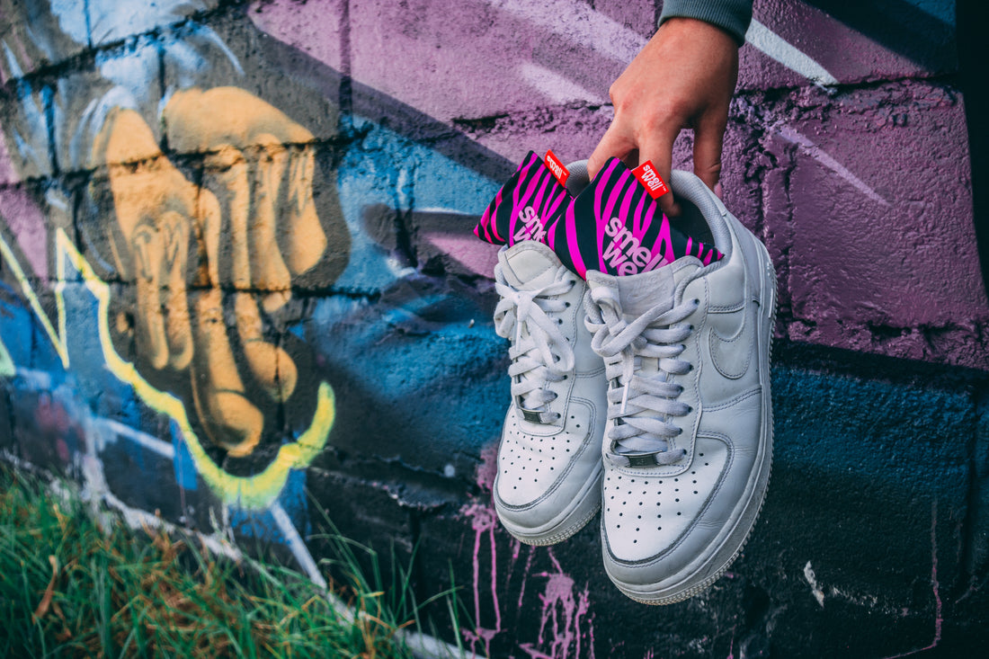A hand holding a pair of white Nike sneakers with pink SmellWell inserts in them infront of a graffiti wall 