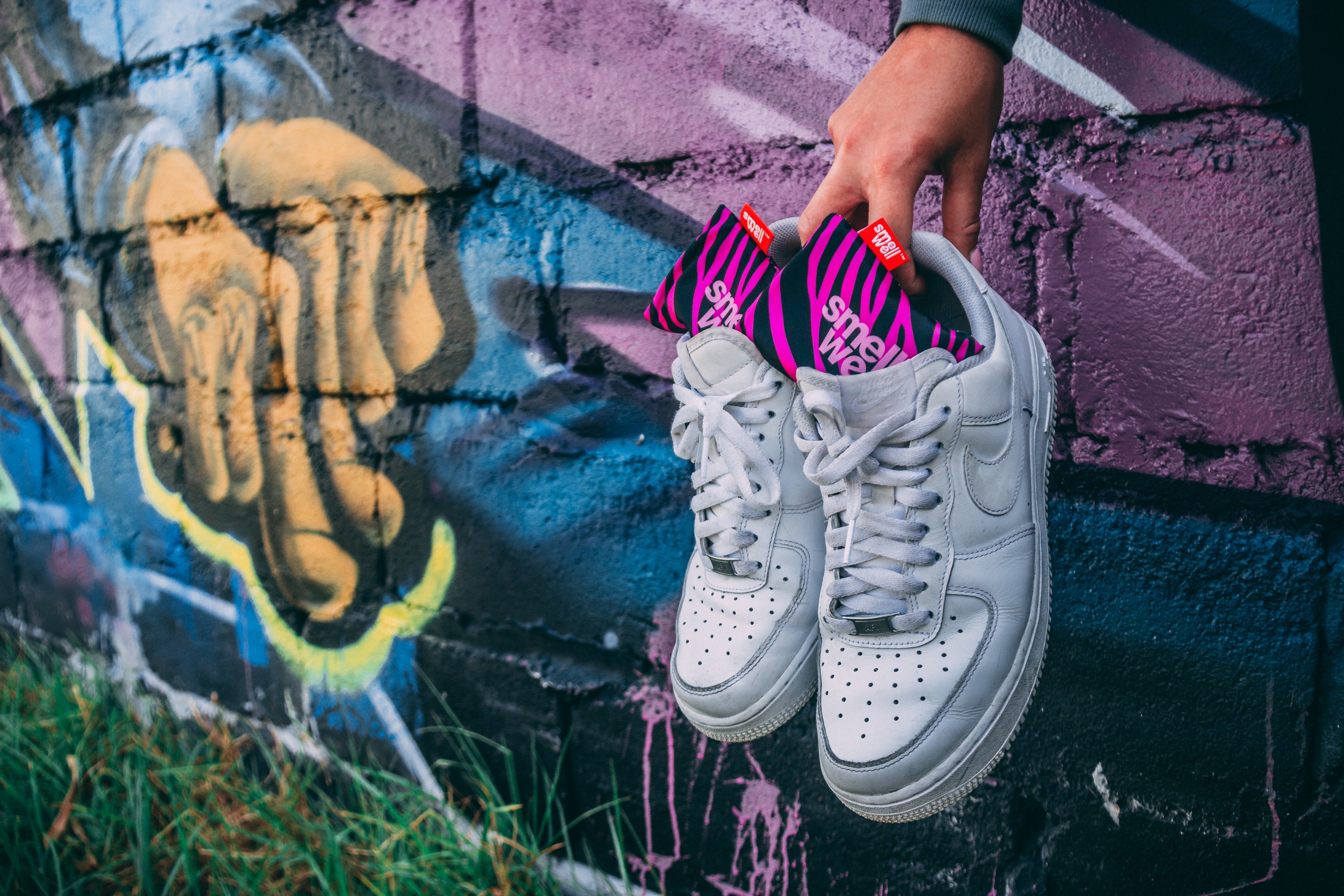 A hand holding a pair of white Nike sneakers with pink SmellWell inserts in them infront of a graffiti wall 