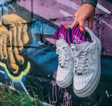 A hand holding a pair of white Nike sneakers with pink SmellWell inserts in them infront of a graffiti wall 